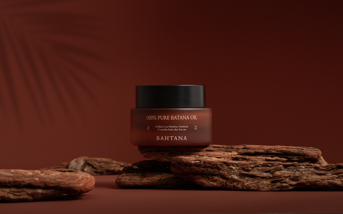 A jar of BAHTANA raw batana oil on bark under palm shadow